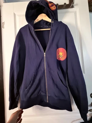 Mörkblå hoodie Bel-Air - Mörkblå hoodie med dragkedja och huva. På bröstet finns ett tryckt emblem i rött och gult med sköld och lagerkrans. Hoodien har kängurufickor och ribbade muddar. Perfekt för en avslappnad och sportig stil. Bel-Air athletic 
