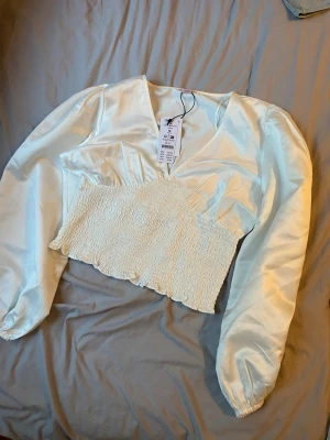 Vit croppad blus från NA-KD - Superfin vit croppad blus från NA-KD med långärmade puffärmar och v-ringning. Toppen har en smockad midja och är gjord i glansigt satinliknande material. Perfekt för att lyfta vilken outfit som helst!