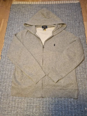 Grå hoodie från Polo Ralph Lauren - Klassisk grå hoodie med dragkedja från Polo Ralph Lauren. Tröjan har huva, ribbade muddar och en liten mörkblå logga broderad på bröstet. Passar dig som är 160 cm lång ungefär och säljer för jag växte ur den