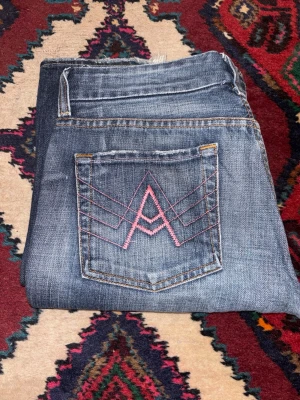 Bootcut jeans från 7 For All Mankind - Snygga blå bootcut jeans från 7 For All Mankind med rosa broderade detaljer på bakfickorna. Jeansen har klassisk femficksdesign, låg midja och slitna kanter vid bensluten. Materialet är jeans med en tvättad look och mjuk känsla. Har ett hål vid grenen.