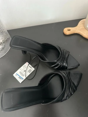 Svarta pumps med klack från Bershka - Säljer ett par svarta pumps från Bershka med öppen tå, fyrkantig tåform och trendig klack. Skorna har en bred rem med snygga veck och är tillverkade i glansigt konstläder. Perfekta för att lyfta din outfit.