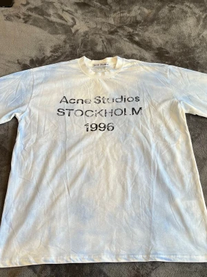Acne Studios t-shirt med tryck dust white/blue - Vit t-shirt från Acne Studios med svart tryck på bröstet där det står 'Acne Studios STOCKHOLM 1996'. Klassisk passform och rund halsringning. Perfekt för en stilren och avslappnad look.