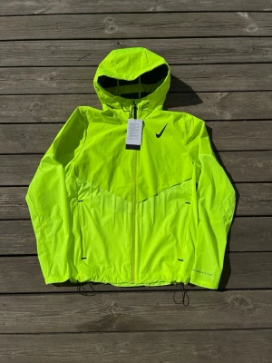 Nike Aeroswift Windbreaker - Snygg neongul vindjacka från Nike med huva och hel dragkedja framtill. Jackan har justerbar dragsko i nederkant och två fickor med dragkedja. Perfekt för löpning eller cykling när du vill synas. Lätt och smidig modell i funktionsmaterial. Nypris 2599kr