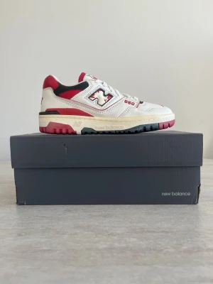 New Balance 550 vita/röda sneakers - Snygga New Balance 550 sneakers i vitt läder med röda och svarta detaljer. Klassisk siluett med lågt skaft, perforerade paneler och tydlig branding på sidan. Yttersulan är i svart och rött gummi för extra kontrast. Perfekta för en sportig och trendig look.