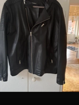 Svart skinnjacka från Produkt - Snygg svart skinnjacka från Produkt med asymmetrisk dragkedja, quiltade detaljer på axlarna och dragkedjor vid ärmslut och fickor. Klassisk bikerstil som ger en cool look till din outfit.