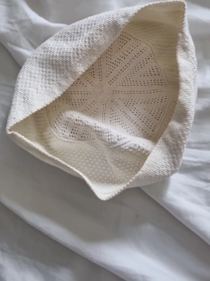 Vit virkad bucket hat - Säljer en vit bucket hat i virkad bomull med snyggt mönster och mjuk känsla. Hatten har en avslappnad form och passar perfekt för en trendig look. Perfekt accessoar för soliga dagar.