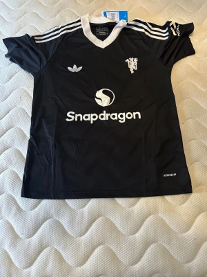 Svart Adidas fotbollströja M - Svart fotbollströja från Adidas med vita detaljer, v-ringad krage och klassiska ränder på axlarna. Tryck med Snapdragon-logga och Manchester United-inspirerad emblem på bröstet. Tillverkad i lätt och ventilerande material, perfekt för träning eller match.