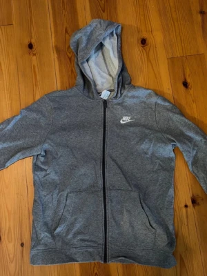 Grå zip-hoodie från Nike - Klassisk grå hoodie från Nike med dragkedja framtill och vit logga på bröstet. Tröjan har huva med dragsko och två stora fickor framtill. Mjuk bomullsblandning som är skön att ha på sig. Perfekt för en avslappnad stil.