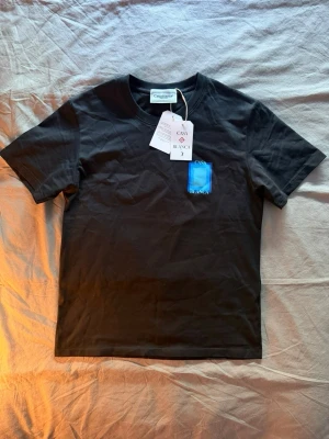 Svart t-shirt från Casablanca - Svart t-shirt från Casablanca med blått grafiskt tryck och texten 'Casa Blanca' på ryggen. Liten blå detalj på bröstet. Klassisk passform och rund halsringning. Perfekt för en stilren och modern look.