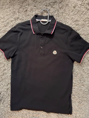Svart Moncler pikétröja med röd/vit kant - Klassisk svart pikétröja från Moncler med vit och röd kant på krage och ärmslut. Tröjan har knappslå framtill och Moncler-logga broderad på bröstet. Perfekt till jeans eller shorts för en clean look.