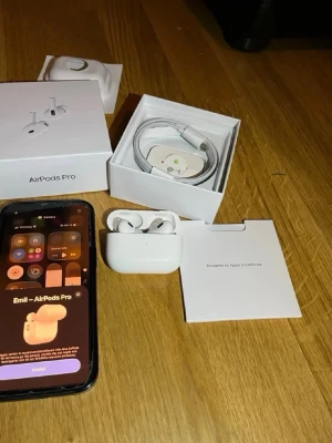 Apple AirPods Pro trådlösa hörlurar - Säljer ett par vita Apple AirPods Pro med laddningsetui och originalförpackning. Hörlurarna är in-ear och har en stilren, kompakt design i blank plast. Medföljer laddningskabel och extra öronproppar. Perfekta för musik, samtal och träning.