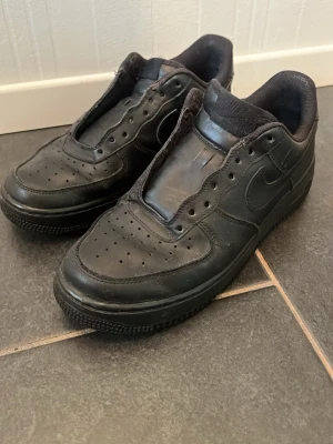 Svarta Nike Air Force 1 sneakers - Klassiska helsvarta Nike Air Force 1 sneakers i läder med perforerad tå och ikonisk swoosh på sidan. Skorna har låg profil och tjock sula med cirkulärt mönster undertill. Perfekta för en clean och stilren streetlook.
