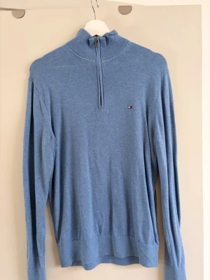 Blå halvzip-tröja Tommy Hilfiger - Snygg blå långärmad tröja från Tommy Hilfiger med halvzip och liten logga på bröstet. Tröjan har ribbade muddar och hög krage. Perfekt för lager-på-lager och enkel att matcha med jeans eller chinos.