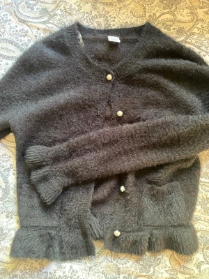 Svart kofta från Lindex - Mysig svart kofta från Lindex med fluffig textur och pärlemorknappar. Koftan har två fickor framtill och ribbade kanter vid ärmslut och nederkant. Perfekt för lager-på-lager och en chill look.