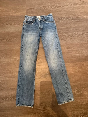 Blå raka jeans med vintage tvätt - Säljer ett par blå jeans med rak passform och klassisk vintage tvätt. Jeansen har fem fickor, knapp och dragkedja framtill samt slitningar som ger en cool look. Perfekta för dig som gillar en avslappnad och tidlös stil.