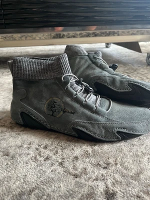Grå höga sneakers med snörning - Unika grå sneakers med högt skaft och snörning. Skorna har en cool design med stickad mudd runt ankeln och detaljer i mocka. Sidan pryds av ett tryck med drakmotiv och texten 'Fashion Sport'. Perfekta för dig som vill sticka ut med din stil.