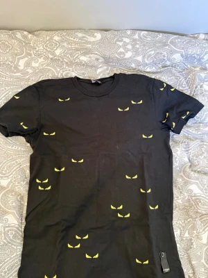 Svart t-shirt med gula ögon från Fendi - Cool svart t-shirt från Fendi med unikt tryck av gula ögon över hela tröjan. Klassisk passform med rund hals och korta ärmar. Tillverkad i 100% bomull för en skön känsla och snygg look.