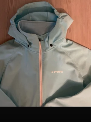 Ljusblå vindjacka från Everest - Snygg ljusblå vindjacka från Everest med dragkedja framtill och två sidofickor med dragkedja. Jackan har huva, hög krage och elastiska muddar vid ärmsluten. Perfekt för blåsiga dagar och outdoor-aktiviteter.