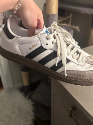 Adidas Samba vita och svarta sneakers - Klassiska Adidas Samba sneakers i vitt läder med svarta ränder och detaljer, samt grå mocka vid tån. Gummisula i brunt och guldfärgad SAMBA-text på sidan. Perfekta för en sportig och stilren look. Nya sko snören med följer! Väldigt fint skick!!!