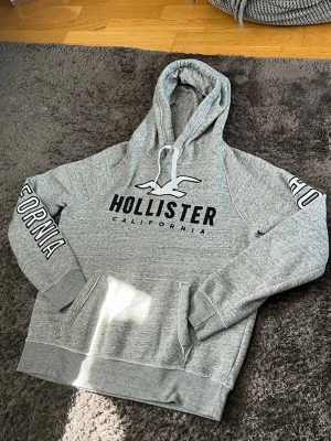 Grå Hollister hoodie  - Snygg grå hoodie från Hollister med stor logga och texten 'California' på bröstet. Tryck på båda ärmarna, känguruficka och dragsko i huvan. Mjuk bomullsblandning som är perfekt för chill dagar.