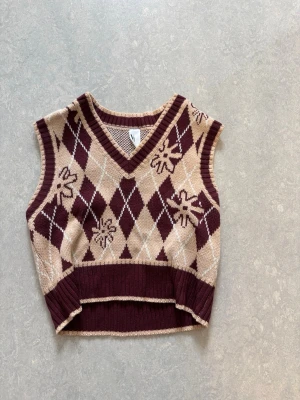 Beige och vinröd stickad väst - Stickad väst i beige och vinrött med klassiskt rutig argyle-mönster och inslag av blommor. V-ringad och croppad modell med ribbade kanter. Perfekt för lager-på-lager och en trendig look.