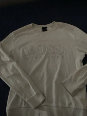 Vit sweatshirt från Hugo Boss - Vit sweatshirt med rund hals och långa ärmar från Hugo Boss. Tröjan har ett diskret broderat BOSS-logo framtill och är tillverkad i mjukt material. Perfekt för en stilren och avslappnad look.