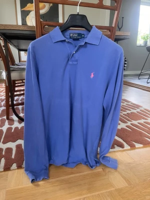 Blå långärmad piké från Ralph Lauren - Snygg blå långärmad pikétröja från Ralph Lauren med klassisk krage och rosa broderad logga på bröstet. Skjortan har knappar vid halsen och är tillverkad i mjuk bomull. Perfekt för en stilren och avslappnad look.