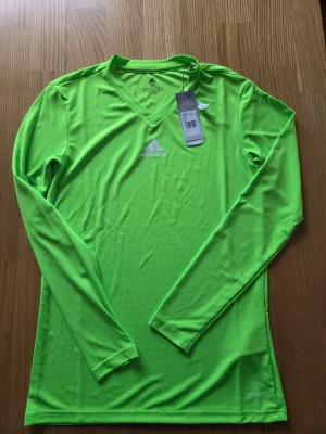 Adidas neongrön långärmad tröja, storlek M - Långärmad tröja från Adidas i en klar grön färg. Tröjan har en V-ringning och Adidas-logotypen är placerad centralt på bröstet. AEROREADY-teknologin är synlig nedtill på tröjan. Tröjan är märkt med "PRIMEGREEN" vilket indikerar att den är tillverkad av återvunnet material. Originaletiketterna sitter kvar på tröjan.