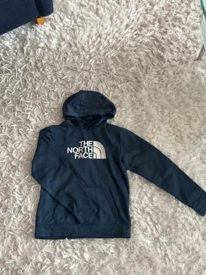 Mörkblå hoodie från The North Face - Snygg mörkblå hoodie från The North Face med stor vit logga på bröstet. Klassisk modell med huva och känguruficka framtill. Tillverkad i mjukt och bekvämt material som passar perfekt till vardagslooken.