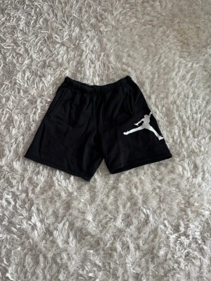 Svarta Jordan shorts med logga - Svarta shorts från Jordan med klassisk vit Jumpman-logga på ena benet. Modellen har elastisk midja med snörning och är tillverkad i mjuk bomull, perfekt för en sportig och avslappnad stil.