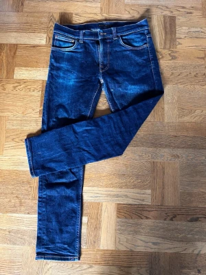 Nudie jeans  - Snygga mörkblå slim fit jeans med klassisk femficksdesign och orangea sömmar. Jeansen har en liten sliten detalj vid ena knät för en cool look. Tillverkade i stretchig denim för extra komfort.