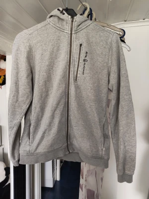 Ljusgrå hoodie Sail Racing - Ljusgrå hoodie från Sail Racing med dragkedja framtill och en bröstficka med logga. Tröjan har huva, ribbade muddar och två sidofickor. Perfekt för en avslappnad och sportig stil. Materialet känns mjukt och bekvämt. Finns hål finns fläckar 