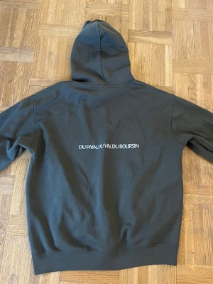 Olivgrön Boursin hoodie med tryck - Säljer en olivgrön hoodie från Boursin med vit text på ryggen 'DU PAIN, DU VIN, DU BOURSIN' och liten Boursin-logga på bröstet. Hoodien har klassisk känguruficka och huva. Perfekt för dig som gillar streetwear och vill sticka ut med ett unikt tryck.