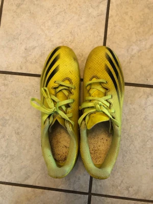 Gula Adidas fotbollsskor med svarta ränder - Säljer ett par gula Adidas fotbollsskor med klassiska svarta ränder på sidan och mönstrad ovansida. Skorna har snörning och dobbar för bra grepp på planen. Tillverkade i syntetmaterial och designade för fart och kontroll på konstgräs.