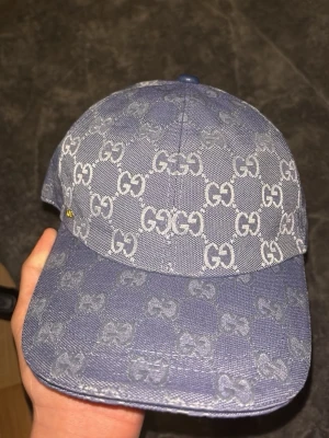 Blå GG-mönstrad keps från Gucci - Snygg blå keps från Gucci med klassiskt GG-monogram över hela kepsen. Tillverkad i denimliknande tyg med justerbart spänne bak i blått läder. Perfekt accessoar för att lyfta din streetwear-look.