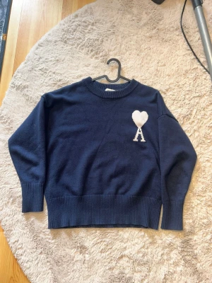Mörkblå stickad tröja Ami Paris - Säljer en mörkblå stickad tröja från Ami Paris med deras ikoniska vita hjärta och bokstaven A broderat på bröstet. Tröjan har rund halsringning, ribbade muddar och nederkant. Perfekt för dig som gillar stilrena och trendiga plagg.