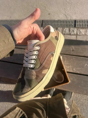 Camo sneakers från Saint Laurent - Säljer ett par snygga Saint Laurent sneakers i camomönstrad canvas med vita snören och beige sula. Klassisk låg modell med gummisula och diskret logga på plösen. Perfekta för dig som vill sticka ut med en streetwear vibe.