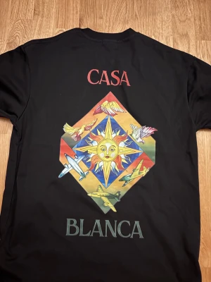 Svart Casablanca t-shirt med tryck - Svart t-shirt från Casablanca med färgglatt grafiskt tryck av sol, fåglar och flygplan på både bröstet och ryggen. T-shirten har rund hals och korta ärmar. Perfekt för dig som gillar unika och kreativa prints.