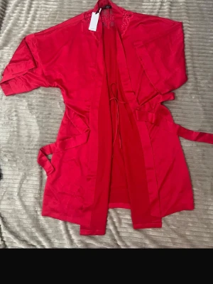 Röd kimono Charlize från Change L - Snygg röd kimono från Change i modellen Charlize. Tillverkad i ett glansigt material med spetsdetaljer vid axlarna och knytband i midjan. Perfekt för att chilla hemma i eller som morgonrock. Färgen är intensivt röd och materialet känns mjukt mot huden.