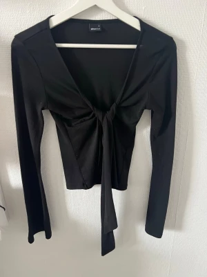 Sommar topp - Trendig svart omlottopp från Gina Tricot med långa ärmar och snygg knytning framtill. Toppen har en djup v-ringning och är tillverkad i ett mjukt, stretchigt material som sitter snyggt på kroppen.