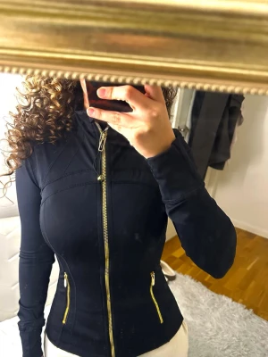 Svart träningsjacka från Lululemon - Snygg svart träningsjacka från Lululemon med guldiga dragkedjor framtill och vid fickorna. Jackan har figurnära passform, hög krage och långa ärmar. Perfekt för gymmet eller löprundan. Materialet är stretchigt och följsamt.