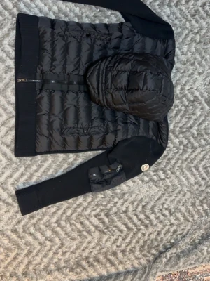Svart pufferjacka från Moncler - Svart pufferjacka från Moncler med quiltad kropp och huva, samt stickade ärmar och muddar. Jackan har dragkedja framtill, två fickor med lock och en ikonisk Moncler-logga på ena ärmen. Perfekt för kalla vinterdagar.