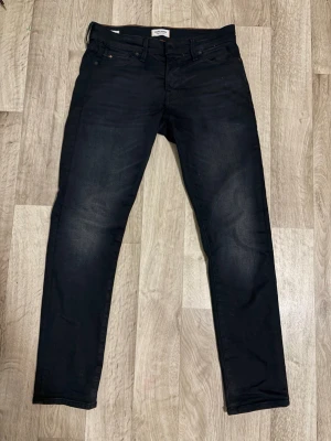 Svarta slim fit jeans från Jack and Jones  - Snygga svarta slim fit jeans från Jack and Jones med klassisk femficksdesign och subtila slitningar på låren. Jeansen har en mörk tvätt och raka ben, perfekta för en stilren och modern look. Materialet är bomull med stretch för extra komfort.