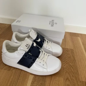 Valentino Garavani vita sneakers - Snygga vita sneakers från Valentino Garavani med breda mörkblå detaljer och nitar över snörningen. Skorna har en klassisk låg siluett och är tillverkade i slätt läder med vita skosnören. Perfekta för dig som vill ha en stilren men ändå unik look.