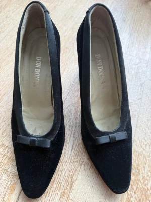 Svarta sammets pumps med rosett från Don Donna - Stilrena svarta pumps från Don Donna i sammet med klack och söt rosettdetalj framtill. Skorna har en klassisk, spetsig tå och en mjuk innersula i beige.  storleken är 39,5 