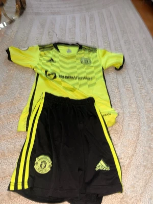 Manchester United träningsset Adidas - Säljer ett Manchester United träningsset från Adidas. Tröjan är gul med svarta detaljer och klubbmärke, och shortsen är svarta med gula ränder och klubbmärke. Materialet är lätt och andas, perfekt för fotbollsträning eller match.