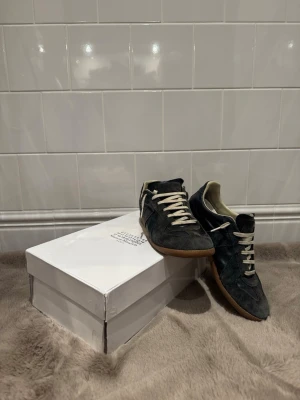 Maison Margiela svarta sneakers - Svarta unika sneakers från Maison Margiela med klassisk design, mocka- och läderdetaljer samt vita snören. Skorna har en låg siluett och gummisula i brunt. Perfekta för dig som gillar stilrena och tidlösa sneakers med en lyxig touch.