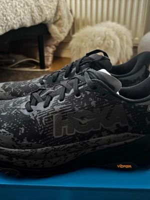  Hoka Speedgoat 6 Gtx  - Snygga svarta sneakers trailskor från Hoka med diskret kamouflagemönster och Vibram-sula. Skorna har lågt skaft, svarta skosnören och en robust, bekväm sula som passar perfekt till streetwear. Materialet är slitstarkt textil och syntet och Gore-tex membran.