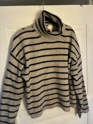 Beige/grå polotröja med svarta ränder H&M - Stickad beige/grå polotröja från H&M med svarta horisontella ränder. Tröjan har ribbad struktur, hög polokrage och långa ärmar. Perfekt för kalla dagar och enkel att styla till olika outfits.