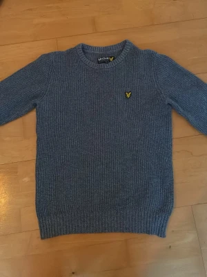 Blå stickad tröja från Lyle & Scott - Säljer en blå stickad tröja från Lyle & Scott med rund halsringning och ribbade muddar. Tröjan har det klassiska gula logomärket broderat på bröstet och är tillverkad i mjukt material. Perfekt för dig som gillar stilrena och bekväma plagg.